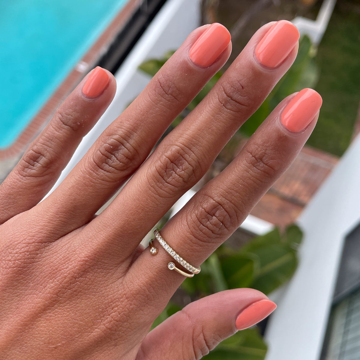 Hand with rounded nails coated in glossy Melon gel polish showing rings near poolside on a sunny day